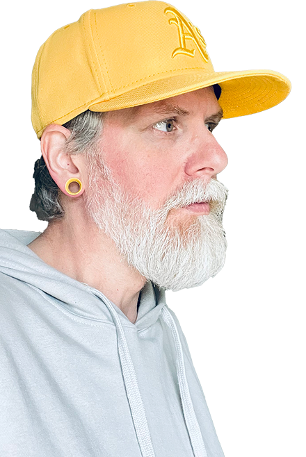 Photograph of James looking coyly at the camera, he’s wearing a light grey hoodie, a purple Oakland Athletics baseball cap and matching 1/2 inch gauge tunnels. His beard is well groomed and grey and white.