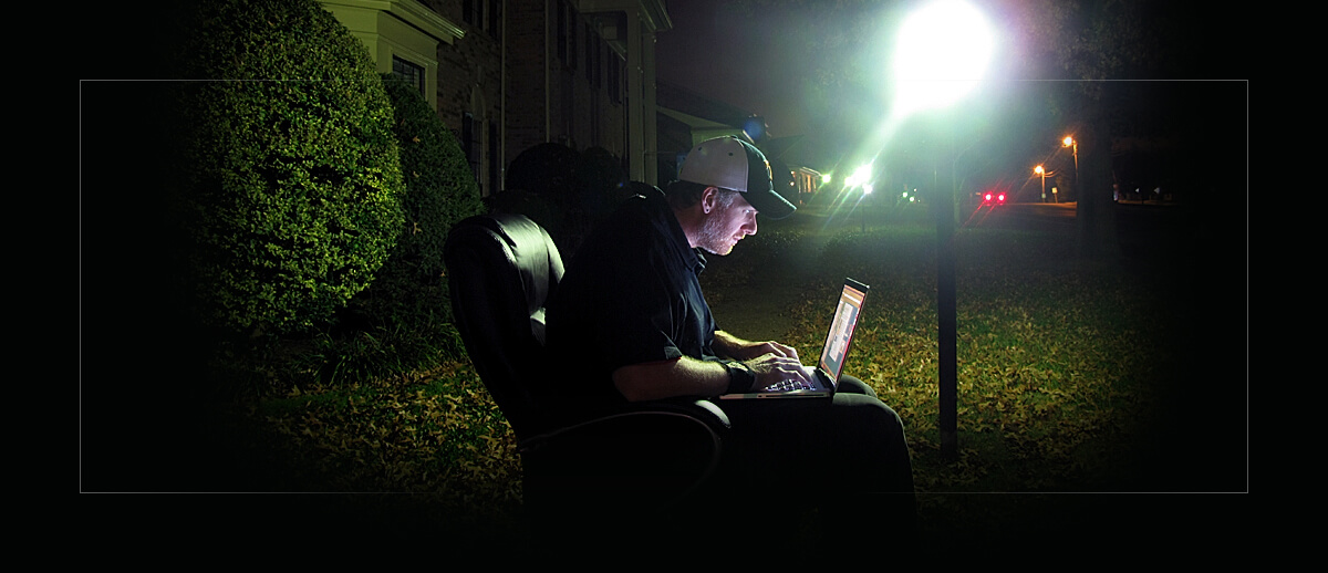 Photograph of a younger James doing work by street light, the screen is vivid but dark. A bright street light illuminates James typing on a laptop while sitting in an office chair outside surrounded by greenery